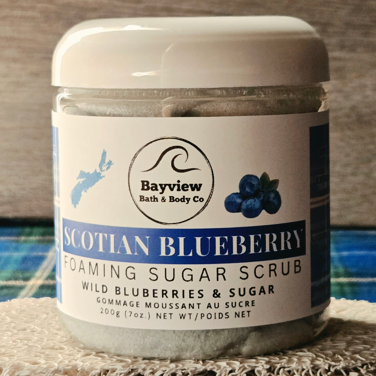 A jar of Bayview Bath & Body Co. Scotian Blueberry Foaming Sugar Scrub, showing the white label with blueberry graphics and the gray scrub inside, set on a textured white cloth.