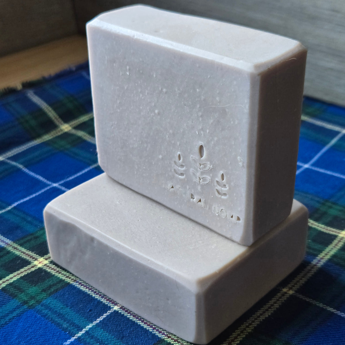 Two rectangular handmade soap bars stacked on top of each other, resting on a blue and green plaid fabric surface.