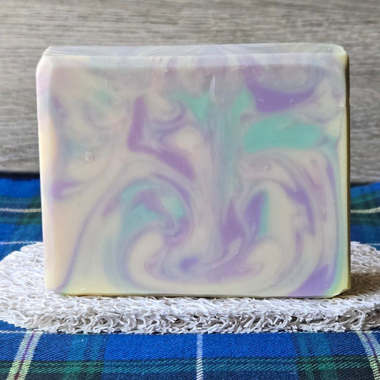 A rectangular bar of handmade soap with a marbled swirl pattern of purple, mint green, and cream, resting on a white lace doily over a blue plaid blanket.