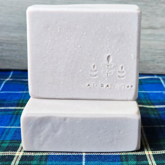 Two rectangular bars of pale lavender soap stacked on top of each other, with the top bar embossed with a leaf design and the words 'NATURAL SOAP'. The soap is placed on a blue and green plaid fabric surface against a gray wooden background.