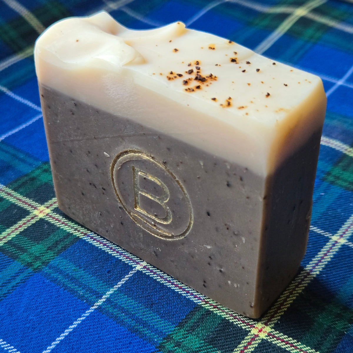 A rectangular bar of soap with a speckled dark gray-brown body and a creamy off-white top layer, featuring a gold embossed letter 'B' and small dark flecks, resting on a blue and green plaid fabric background.